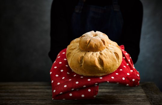 Food photograph of a woman in a black jumper and denim apron holding a homemade loaf of cottage bread showing its distinctive shape, the loaf is sat on a red and white polka dot patterned tea towel