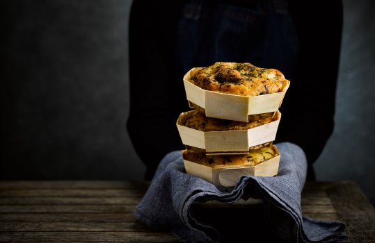 Food photograph of a woman in a black jumper and denim apron holding three miniature homemade focaccia’s, the breads are stacked on top of each other in wooden baking baskets, and sat on a denim tea towel