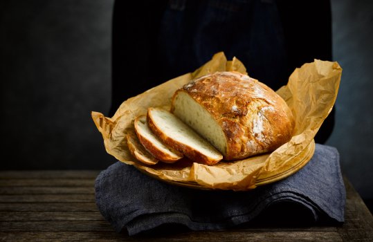 Food photograph of a woman in a black jumper and denim apron holding a homemade loaf of no knead bread, the loaf is sat on a denim tea towel and lined with brown baking paper, three slices have been taken from the front of the loaf to show the crumb structure