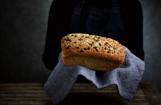 Food photograph of a woman in a black jumper and denim apron holding a homemade loaf of seeded wholemeal bread, the loaf is sat on a denim tea towel