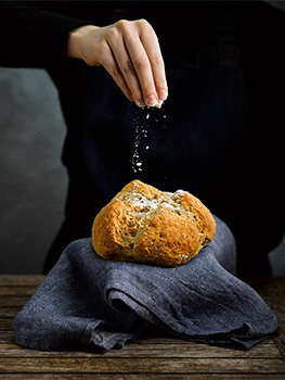 Food photograph of a woman in a black jumper and denim apron holding a homemade loaf of honey and oat soda bread, the loaf is sat on a denim tea towel
