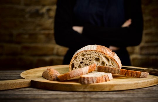 Food photograph of a woman in a black jumper and denim apron with folded arms stood behind a sliced loaf of sourdough bread, with slices removed to show the open crumb structure on the face of the loaf