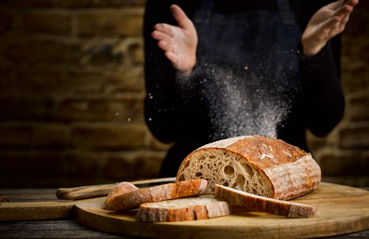 Food photograph of a woman in a black jumper and denim apron, clapping flour off her hands; stood behind a sliced loaf of sourdough bread, with slices removed to show the open crumb structure on the face of the loaf