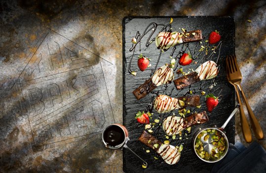 Aerial food photograph of white chocolate dipped brownie sticks sprinkled with pistachio kernels and drizzled with chocolate sauce, shot on a slate against a mottled grey background with an embossed Bakeshed logo
