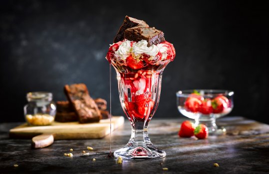 Close up food photograph of an ice cream sundae with strawberries, vanilla ice cream, whipped cream, chopped hazelnuts and chunks of Belgian chocolate brownie on top. shot on a dark set with ingredients in the background
