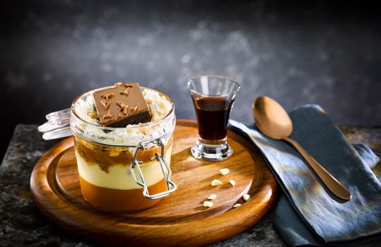 Close up food photograph of a caramel trifle in a kilner jar with custard, caramel sauce, whipped cream, chunks of chocolate caramel brownie on top. shot on a dark set with a shot of coffee liqueur in the background