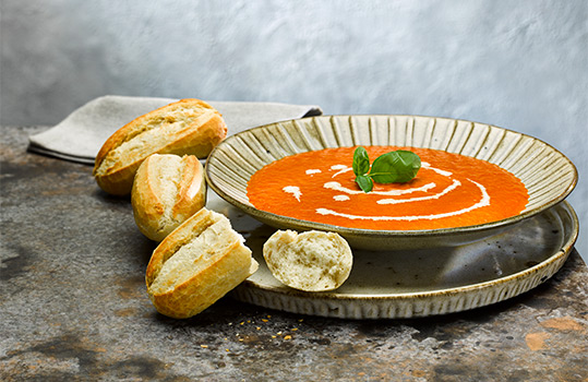 Food photograph close up of a bowl of tomato soup with a swirl of cream, accompanied by several white petit pain rolls, one of which is broken open to show the interior crumb