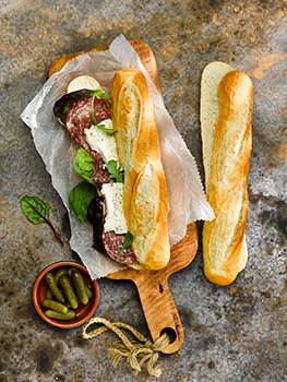 Aerial food photograph of a salami and mozzarella cheese white baguette sandwich, with basil leaves and sliced pickles on a wooden chopping board, shot on a splattered grey background
