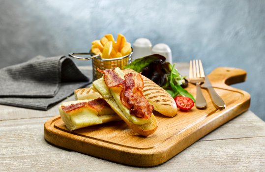 Food photograph of a brie and bacon panini baguette sandwich, shown open faced with the lid of the sandwich on the side, alongside chips and salad on a wooden chopping board, shot on a light grey background