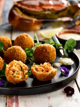 Food photograph of crab arancini, golden crunchy deep fried balls of crab and sweetcorn risotto, served on a black plate with nasturtiums and tartare sauce, on a painted blue and red tabletop with a whole crab in the background