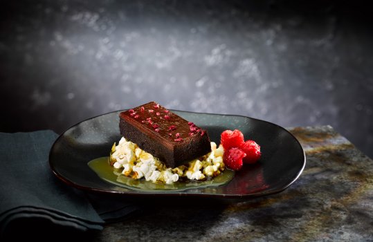 Food photograph of a slice of dark chocolate and raspberry brownie shown on top of a disc of popcorn brittle with caramel sauce, shown on a black plate alongside fresh raspberries