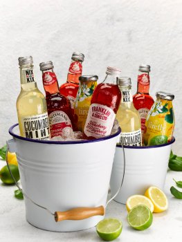 Drinks photograph of two ice buckets of bottled summer soft drinks, served on a white tabletop with wedges of lemon and lime, with scattered basil leaves
