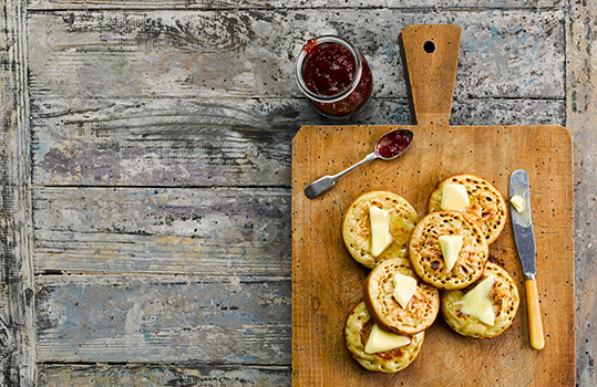 Aerial food photograph of homemade crumpets stacked on a wooden board with a jar and spoon of strawberry jam, and each crumpet topped with a generous pat of butter