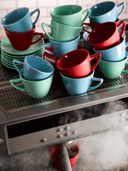 Food photograph of the making of a cup of coffee, stacks of multicoloured coffee cups on top of a commercial coffee machine, one cup in the machine being poured into, with clouds of steam billowing out from the coffee machine