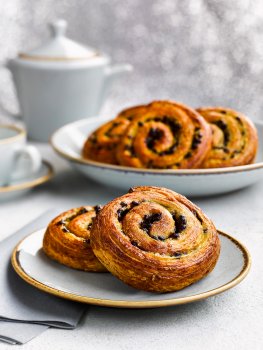 Food photograph close up of danish pastry swirls, two pastries stacked up on a plate with a platter of the same pastries, a teapot and cup and saucer in the background, with a shimmering flittered white background