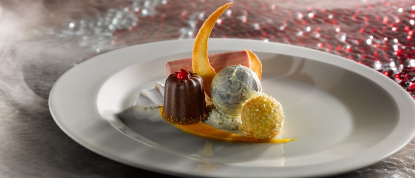 Food photograph of a fine dining dessert, a mango and coconut bonbon, a dark and white chocolate truffle, a raspberry parfait and a canele served with mango sauce on a white plate on a red textured glass background, with clouds of smoke