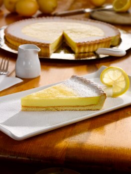Food photograph of a slice of lemon tart on a rectangular white plate, dusted with icing sugar and garnished with a slice of lemon, with the whole tart with a slice taken out in the background, along with a selection of lemons