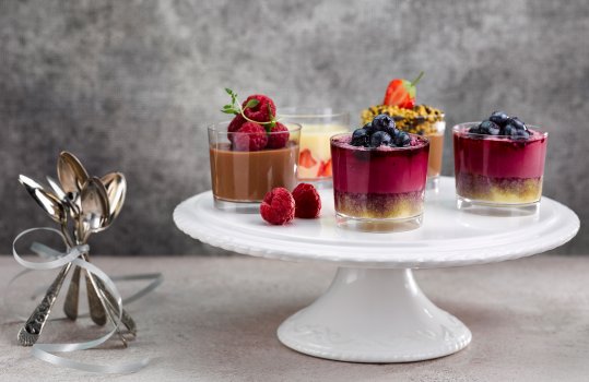 Food photograph of dessert pots, small transparent plastic cups filled with blueberry cheesecake topped with blueberries, white chocolate mousse with strawberries, and chocolate mousse with raspberries, served on a white china cake stand on a grey table along with a bundle of teaspoons