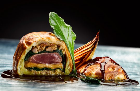 Food photograph close up of a fine dining dish, a pithivier of duck breast and foie gras wrapped in spinach with bacon and onions encased in crisp golden puff pastry served on a sheet of glass with a charred shallot and jus