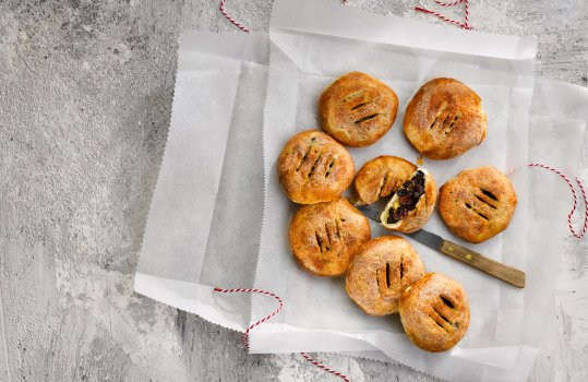 Aerial food photograph of home made Eccles cakes, eight crisp golden brown Eccles cakes served on baking paper with twine, one cake cut in half to show the juicy plump raisin filling, served on a grey concrete tabletop