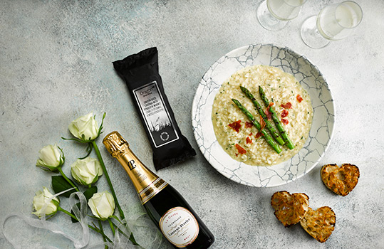 Aerial food photograph of a Valentines Day champagne risotto, with asparagus, crispy prosciutto and heart shaped sourdough croutons. Shot on a light grey stone background with white roses, champagne and Dragon Handcrafted Cheddar with Penderyn Whisky