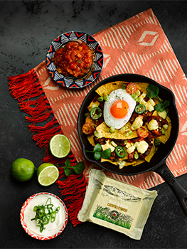 Aerial food photograph of a cast iron skillet filled with homemade chilaquiles, fried corn tortilla chips with black beans, tomato, spring onion, coriander leaves, Dragon Welsh Caerphilly cheese and topped with a fried egg. Shot on a dark slate background with limes, salsa, sour cream and a pack of Dragon Caerphilly Cheese