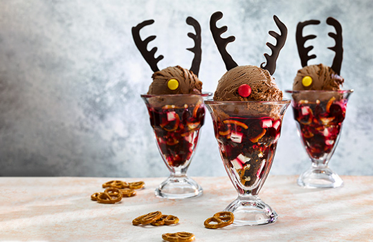 Food photograph of a trio of Christmas ice cream sundaes, chocolate ice cream balls sitting on top of chocolate brownie, pretzel, marshmallow and strawberry sauce in sundae glasses, decorated with tempered chocolate antlers and coloured chocolate noses. One sundae has a red nose while the others are all yellow, like Santa’s reindeers