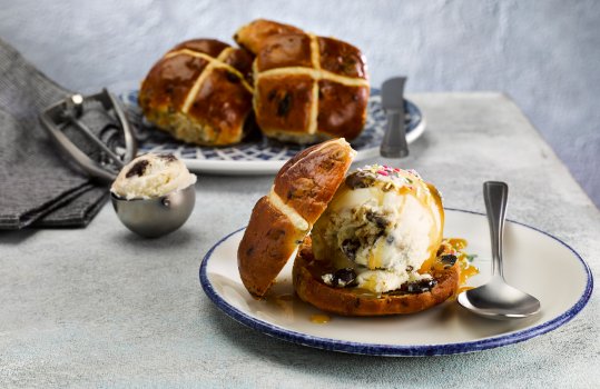 Food photograph of an Easter ice cream sandwich, a toasted hot cross bun, sliced and filled with a large scoop of rum and raisin ice cream and drizzled with caramel sauce, shot on a light grey stone background with more toasted hot cross buns in the background
