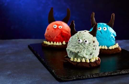 Food photograph close up of three Halloween ice creams, double chocolate cookies topped with colourful balls of ice cream, decorated with white chocolate claws, tempered chocolate horns and fondant eyes. Shot on a slate board against a dark blue background