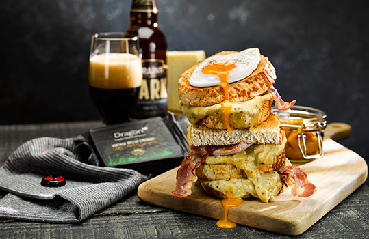 Close up food photograph of a Welsh rarebit bacon and egg toastie stack, thick slices of toasted tiger bread topped with oozing Welsh rarebit, crispy bacon, and a fried egg whose yolk is dripping down the stack onto the board. Shot on a dark wood background alongside a bottle and glass of dark beer and a block of Dragon Vintage Cheddar