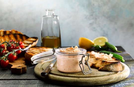 Food photograph of a small swing top jar filled with smoked salmon paté, alongside cherry tomatoes and sourdough toast, one piece of toast is spread with the salmon paté