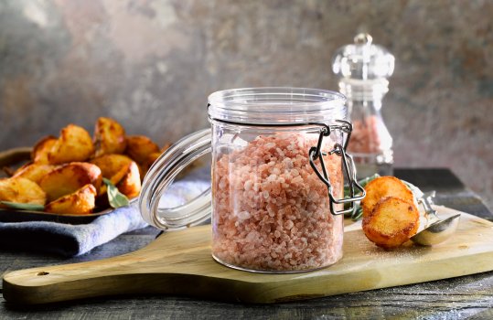 Food photograph of a swing top jar filled with Himalayan pink rock salt, alongside a stack of crispy golden roast potatoes on a wooden cutting board