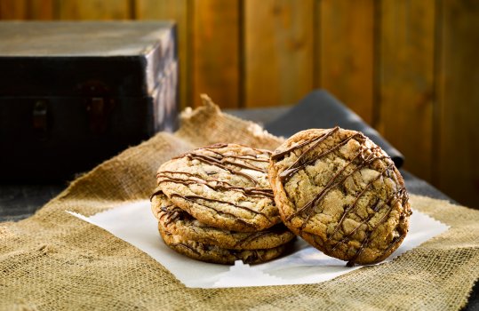 Food photograph close up of four chocolate chip cookies, drizzled with shiny tempered milk chocolate, photographed on wax paper and hessian cloth