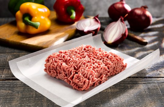 Food photograph close up of a package of raw lamb mince, shot in a dark grey set alongside red onions and peppers