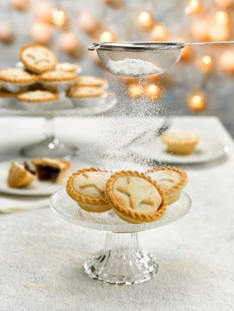 Food photograph of a stack of vegan mince pies being sprinkled with icing sugar from a vintage strainer, shot on a light set with festive fairy lights