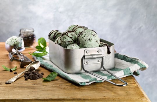 Food photograph of several scoops of mint chocolate chip ice cream served in an army style mess tin, with toppings and sauces on the tabletop in the background