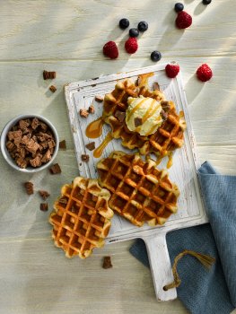 Aerial food photograph of three Belgian style waffles on a white cutting board, one of which has been topped with a scoop of ice cream and drizzled with caramel sauce