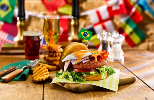 Food photograph of a Brazilian burger in a World Cup themed pub setting with flag bunting, a chorizo patty topped with a fried egg, sliced ham and cheese sauce, with a miniature Brazilian flag stuck in it.