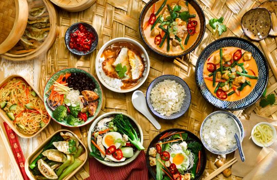 Aerial food photograph of a spread of Japanese dishes, featuring ramen noodle bowls, steamed rice, chicken katsu curry, tofu raisukaree, gyoza and noodle salad. Shot on a large woven palm leaf with chopsticks and steam baskets