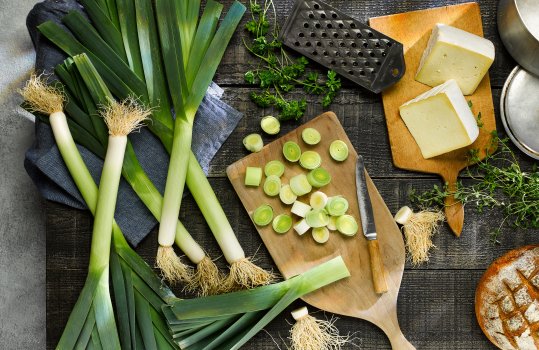 Aerial food photograph of a tabletop with whole Welsh leeks, chopped Welsh leeks on a board, Caerphilly cheese wedges and a loaf of crusty bread; next to an empty saucepan and cheese grater, ready to make leek soup and cheese on toast