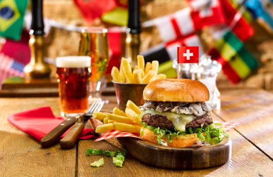 Food photograph of a Swiss burger in a World Cup themed pub setting with flag bunting, a beef patty topped with mushrooms in cream sauce, and Swiss cheese, with a miniature Swiss flag stuck in it.