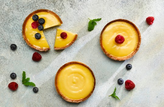 Aerial food photograph of three individual lemon tartlets, shot on a grey stone background with mint leaves, fresh raspberries and fresh blueberries; one tart has a wedge sliced out of it to show the texture of the filling