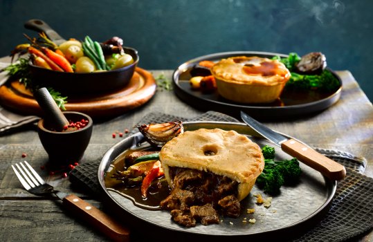 Food Photograph of a gluten free steak pie with roasted vegetables and gravy. The pie is cut open to reveal the pieces of steak in the filling