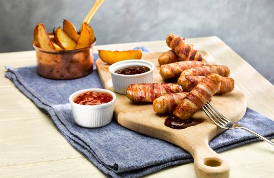 Food photograph of bacon wrapped honey and rosemary chipolata sausages, shot on a wooden serving board with brown sauce, alongside potato wedges