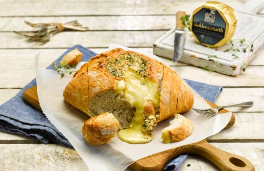 Food photograph of a wheel of Golden Cenarth cheese, baked inside a loaf of garlic bread, the corner of the bread has been torn so that the molten baked cheese flows over the serving board