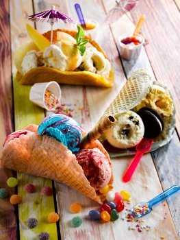 Food photograph of multi coloured ice cream tacos, scoops of bubblegum, strawberry, oreo, caramel and vanilla ice creams served in taco shells made from waffle batter, garnished with jelly tots, jelly beans, wafers, sprinkles, strawberry sauce, oreos and cocktail umbrellas; served on a multi coloured painted wooden table