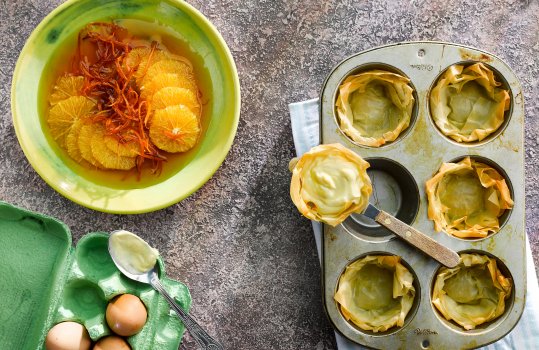 Aerial food photograph showing the making of vanilla custard and orange filo pastry tarts, six crispy filo shells in a muffin tin, one having been removed and filled with custard, shot alongside a box of eggs, and a bowl of orange slices and candied orange peel, marinating in syrup, shot on a grey stone background