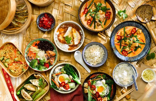 Aerial food photograph of a spread of Japanese dishes, featuring ramen noodle bowls, steamed rice, chicken katsu curry, tofu raisukaree, gyoza and noodle salad. Shot on a large woven palm leaf with chopsticks and steam baskets