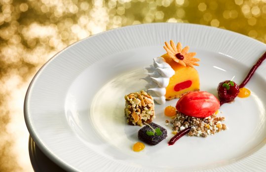 Food photograph of a fine dining dessert, mango and raspberry parfait topped with Italian meringue, an almond brownie and a rocher of raspberry sorbet served on a white plate on a reflective glittering gold background